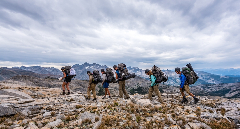 A group of students wearing backpacks high across rocky terrain with mountains in the background. 